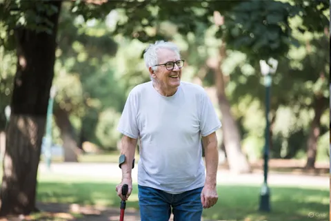 Persona mayor caminando en un parque con bastón, sonriente, representando la recuperación funcional en EAP con ejercicio.